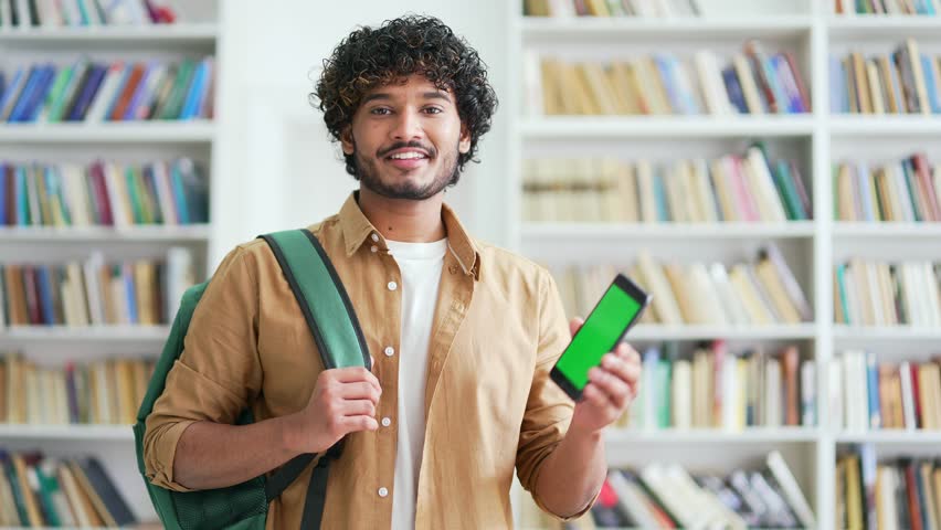 Smiling young university student showing smartphone with green screen while standing in campus library space Color key vertical mockup. Template for advertising. Male shows the phone looking at camera Royalty-Free Stock Footage #1103224451