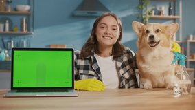Portrait of a Smiling Young Woman in Yellow Rubber Gloves With Her Dog in the Kitchen.Female Housewife with a Laptop with a Green Screen Showing an Okay Sign.Organizing Pet Supplies and Spaces.
