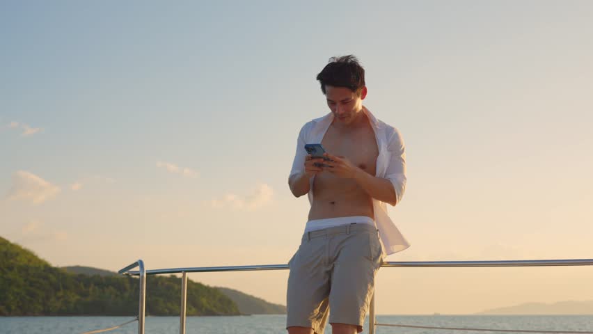 Asian young man tourist using smartphone taking photo during yachting. Attactive male feel happy and relax while sitting alone on deck of yacht, enjoy time when catamaran boat sailing during summer.