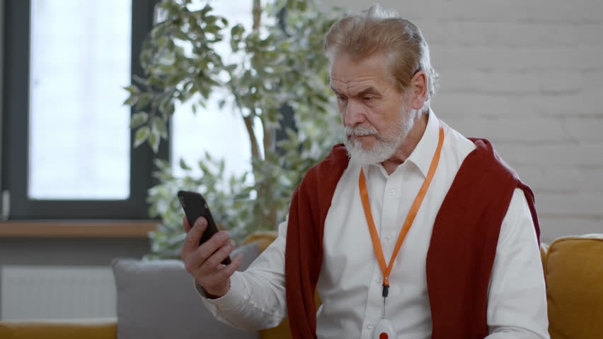 Healthcare help service. Senior man web surfing on smartphone and feeling sudden acute headache, pushing red emergency button on his chest, empty space