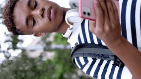 Vertical screen video of young hispanic latin man in striped shirt with headphones holding smart mobile phone standing outdoor looking for address with smartphone app in city street. HD shot footage