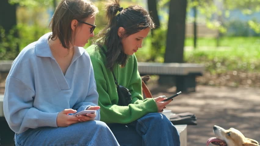 Two Bored friends girls using their phones sitting on bench in the park . High quality FullHD footage Royalty-Free Stock Footage #1103243239