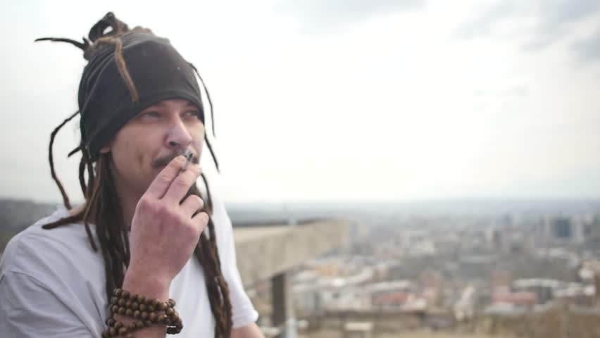 Portrait of one young attractive guy smoking a cigarette with dreadlocks and an earring in his ear and a bracelet on his hand made of balls. Handsome brazilian african man smoking while thinking