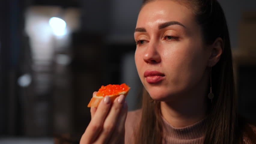 Close-up Caucasian young woman eating sandwich with spoiled salmon caviar making dissatisfied facial expression. Unsatisfied beautiful lady with bad taste toast indoors