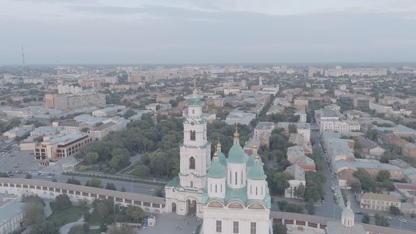 Astrakhan, Russia. The Cathedral of the Assumption of the Blessed Virgin and its adjoining bell tower stand at the heart of the Astrakhan Kremlin, a fortified complex on a hill in the Volga Delta. The cathedral displays five green domes with golden crosses and a tall white bell tower, all enclosed within historic stone walls. The camera pulls back from a close aerial perspective to reveal the full fortress grounds, surrounding gardens, and the dense urban grid of Astrakhan stretching toward the horizon. Soft evening light and a muted sky create a calm atmosphere over the city. The Assumption Cathedral was constructed between 1699 and 1710 and is regarded as a notable example of early eighteenth-century Russian church architecture, Departure of the camera. A-Log.