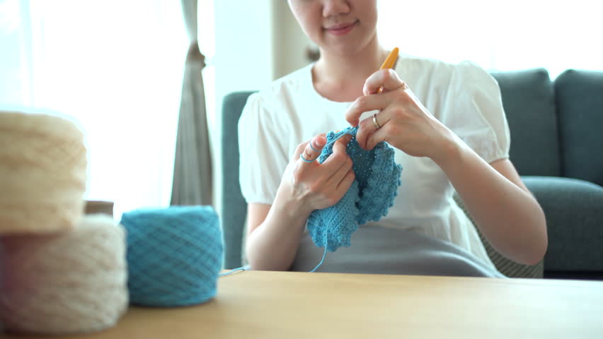 Happy cheerful Asian woman doing a crochet in living room in free time. Skillful Asian craftswoman knitting a handcraft crochet sweater and hat in cozy living room.