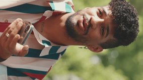 VERTICAL VIDEO: Close-up of young smiling male with curly hair wearing striped shirt sitting in park having picnic on summer day outdoors, talking with friends, using cellphone