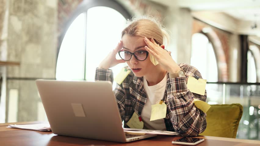 Nervous stressed young blond woman freelancer with stickers typing quickly on laptop computer and looking at paper documents asking to hurry up scream and warning about deadline at coworking workplace