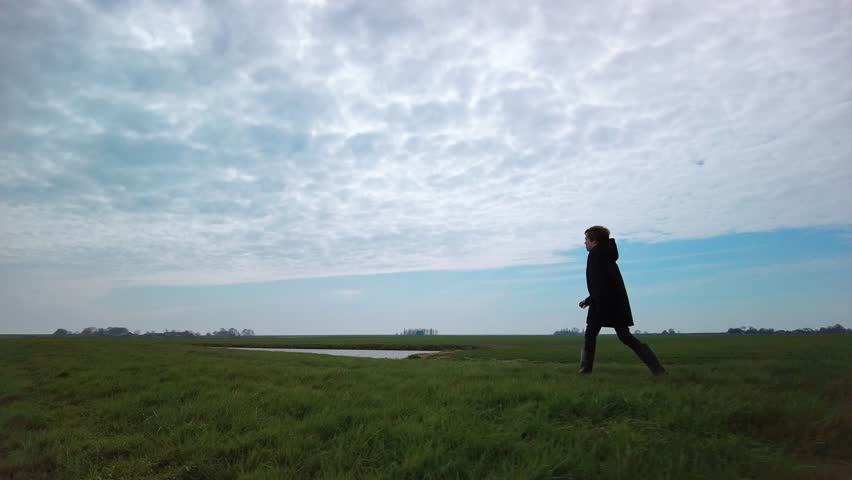 Child in winter coat and boots walks briskly steadfast alone over the green grass trail through the rural landscape and farmlands in early spring on a cloudy day. Low angle left side view slow motion. Royalty-Free Stock Footage #1103282995