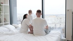 Rear view of loving father kissing his baby daughter while little girl hugging caring mom in morning after waking up. Young married couple with kid resting on white blanket in bedroom of modern flat. - Powered by Shutterstock - Get 15% off with code: PIKWIZARD15