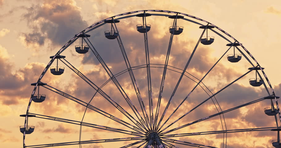 Spinning Carnival Ferris Wheel in amusement park against scenic sunset sky clouds