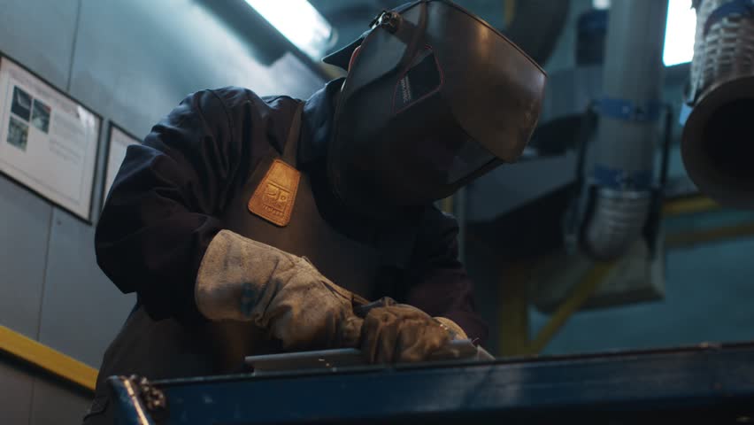 Asian man is focused on work. Uniformed welder lifts protective mask and examines detail. Male is satisfied with result and smiles. Men's labor. Diversity of men's profession. Royalty-Free Stock Footage #1103292531