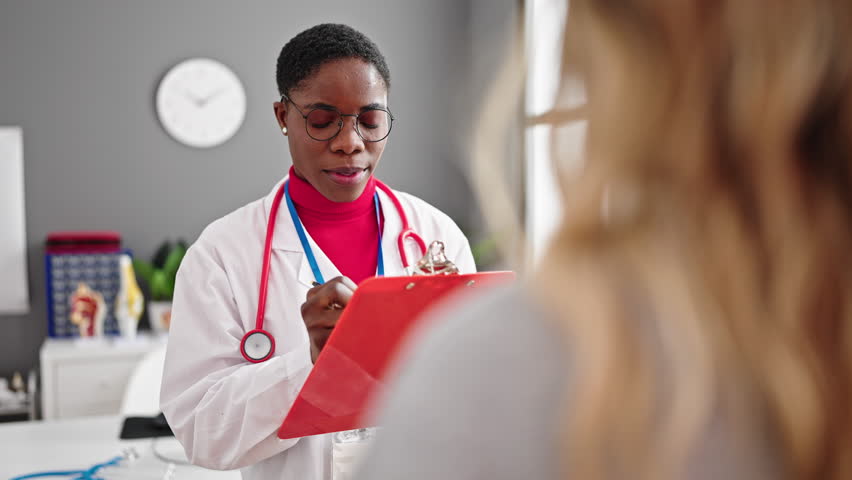 African american woman doctor writing medical report speaking with patient at clinic Royalty-Free Stock Footage #1103295171