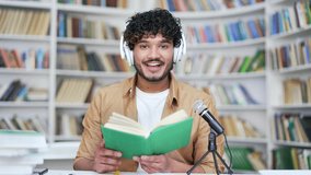 Young college teacher in wireless headphones conducting online training in campus library space. A university tutor with a book reads a lecture or records a podcast with a microphone looking in camera
