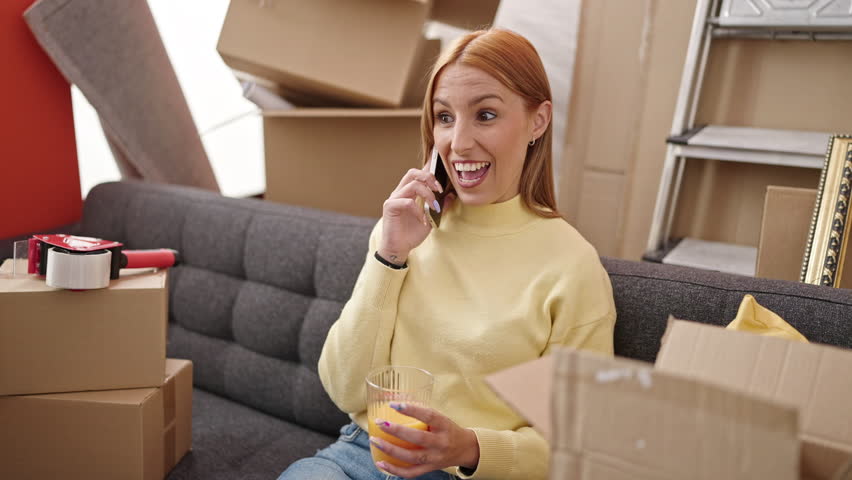 Young blonde woman talking on smartphone drinking orange juice at new home