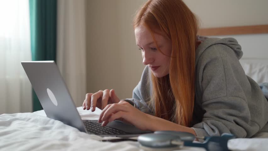 Young woman talking on smartphone while using laptop at home on bed. Teenage girl speaking with friends mobile phone working computer, studying typing, online shopping, watching video, reels Royalty-Free Stock Footage #1103311539
