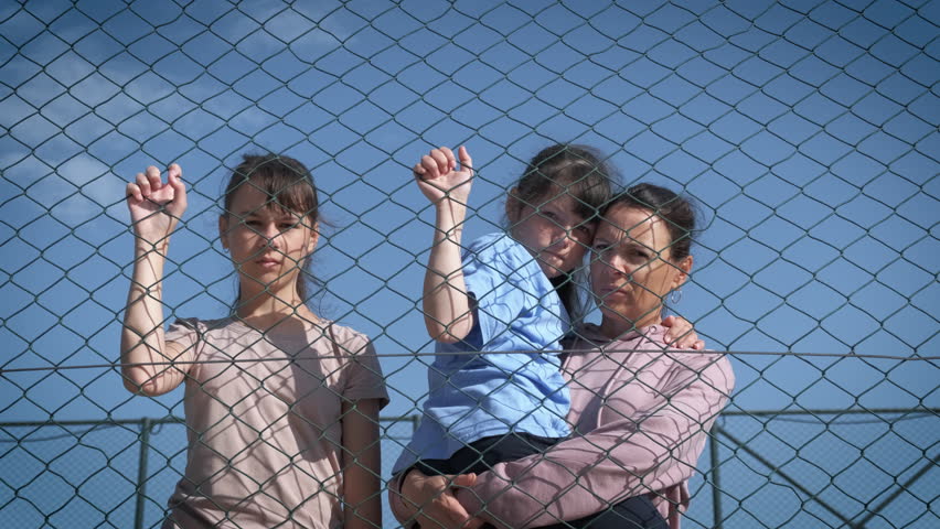 Desperate refugees. A mother with her daughters in despair behind a wire fence look sadly at the camera. The problem of assimilation of refugees. Royalty-Free Stock Footage #1103315809