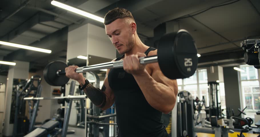 Close-up view of sporty man performing barbell curls to gain lean biceps. Portrait of caucasian sportsman lifting heavy weights in gym studio