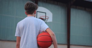 Video of a boy standing on a basketball court and holding a basketball. Cinematic shot of teenager standing with a ball in his hand and a basketball basket behind him