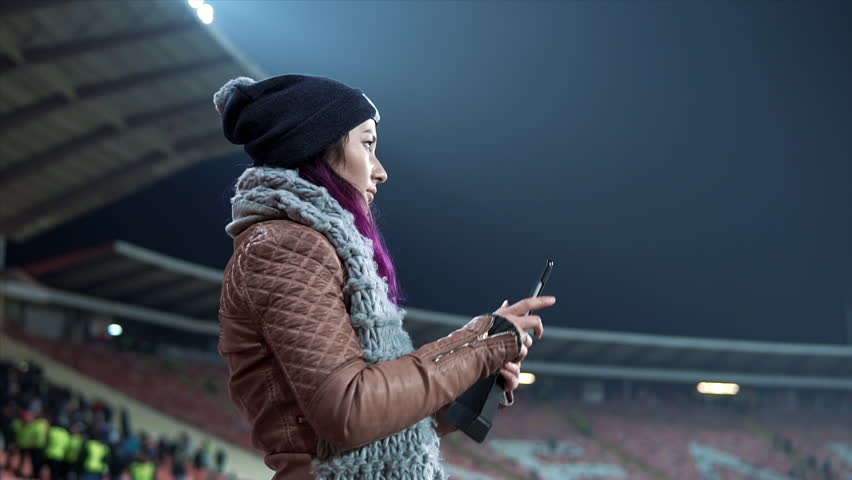 Woman taking photos or recording videos of live music concert with smartphones at open air festival or at sport game. Bright colorful stage lighting. Girl dressed in hat and scarf shooting video at