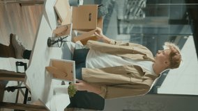 Vertical Video,.Young Upset Male Worker Stands at Table in Bright Office Room.Packs His Workplace Stuff and Flowerpot in Carton Box Because of Dismissal. Leaving The Office.Resignation Concept