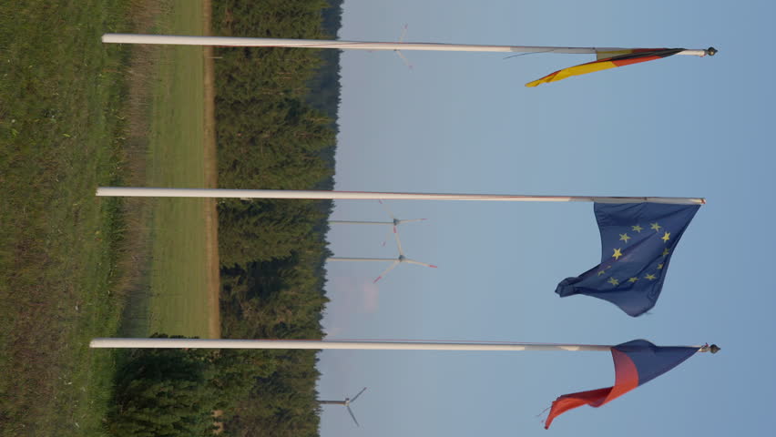 Wide shot vertical video of German, Czech Republic, and European Union flags on flagpoles at background of hills with windmills spinning. Car passing at front of national symbols fluttering in wind Royalty-Free Stock Footage #1103325311