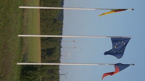 Wide shot vertical video of German, Czech Republic, and European Union flags on flagpoles at background of hills with windmills spinning. Car passing at front of national symbols fluttering in wind - Powered by Shutterstock - Get 15% off with code: PIKWIZARD15