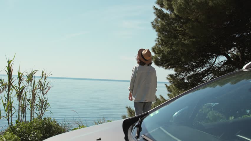 A young Caucasian woman enjoying the top view while standing next to the car during her solo trip to the sea.