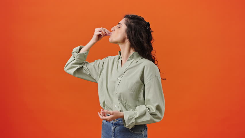Caucasian emotional young curly haired woman in casual clothes, showing tasty gesture, kisses fingers and says bellissimo, looking at camera isolated on orange background. Gourmet culinary concept