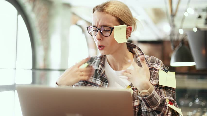 Nervous stressed young blond woman freelancer with stickers typing quickly on laptop computer and looking at paper documents asking to hurry up scream and warning about deadline at coworking workplace