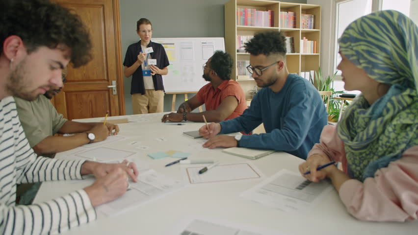 Group of multi-ethnic immigrant students listening to female teacher and taking notes during English lesson in classroom