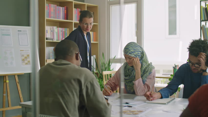 Female teacher walking in classroom and talking to immigrant students as they taking test during lesson in language school
