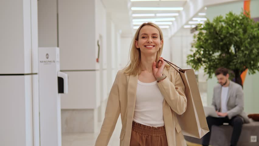 Gorgeous, happy, beautiful, joyful blonde girl holds shopping bags in hands and looking around a shopping center. Woman smiles, enjoys her purchases while people are in the background using gadget.