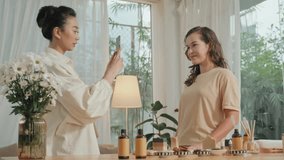 Medium shot of young Asian woman recording video on smartphone of her girlfriend showing how to make handmade soap at home