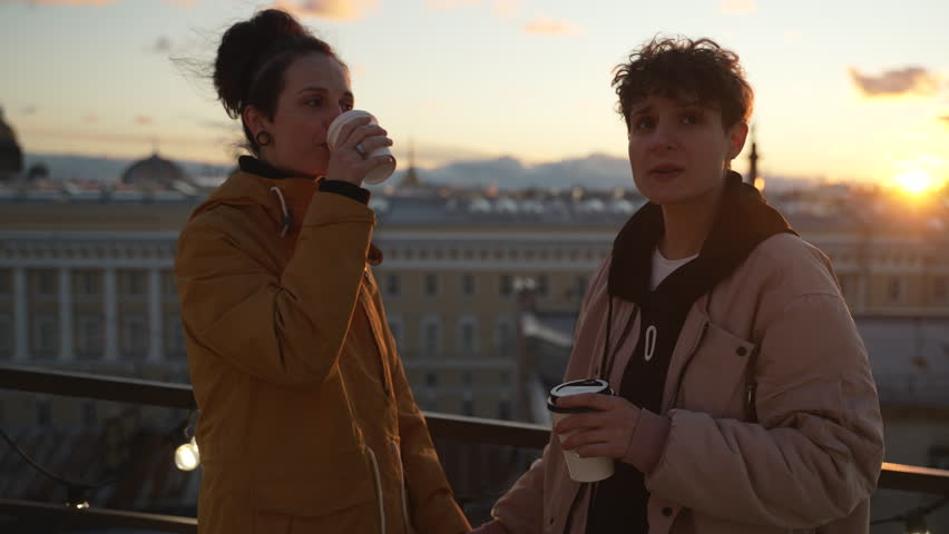 4k Two lgbt women drink coffee on rooftop in city spbd. Young lesbian couple drinking hot drink and looking with smiles, hugging and standing on background of town during sunset. Happy non-binary | Shutterstock HD Video #1103341217