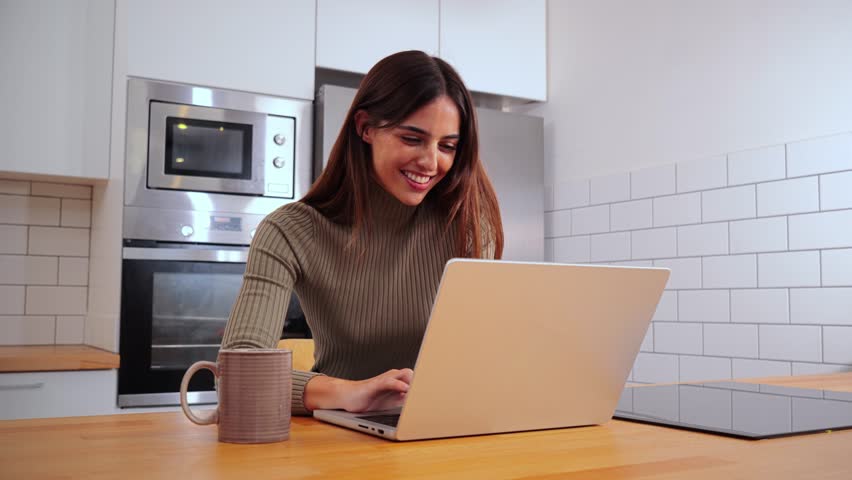 Young entrepreneur woman working online on laptop at home office. Happy young girl studing with a notebook at her apartment kitchen. Positive brunette female using a computer to browse on internet