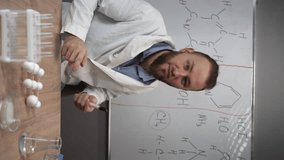 a chemistry teacher or tutor looks at the camera, points to a whiteboard and talks about formulas. vertical video. Video conference webcam portrait