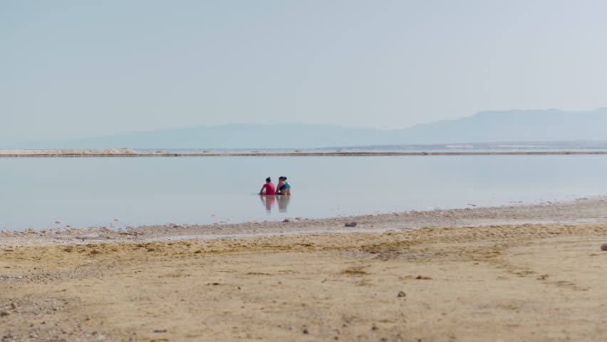 2 girls sitting in the water of the Dead Sea with a spectacular mountainous view - wide shot