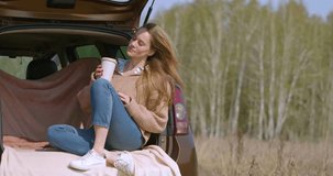 Coffe break in the car trunk after lonely driving. A young woman drinks coffee rest in the trunk of her car in nature. Close-up 4k video.