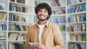 Portrait of young smiling university student holding tablet and looking at camera while standing in campus library space. Happy positive friendly male with backpack in college classroom posing - Powered by Shutterstock - Get 15% off with code: PIKWIZARD15