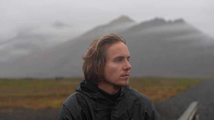 Side view close-up shot of a blond man wearing a raincoat while standing in the middle of a countryside road near a mountain on a cloudy day