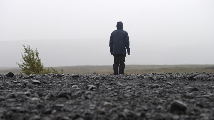Tilting back view whole body shot of a man wearing a raincoat as he stands in the middle of an unpaved road in Iceland