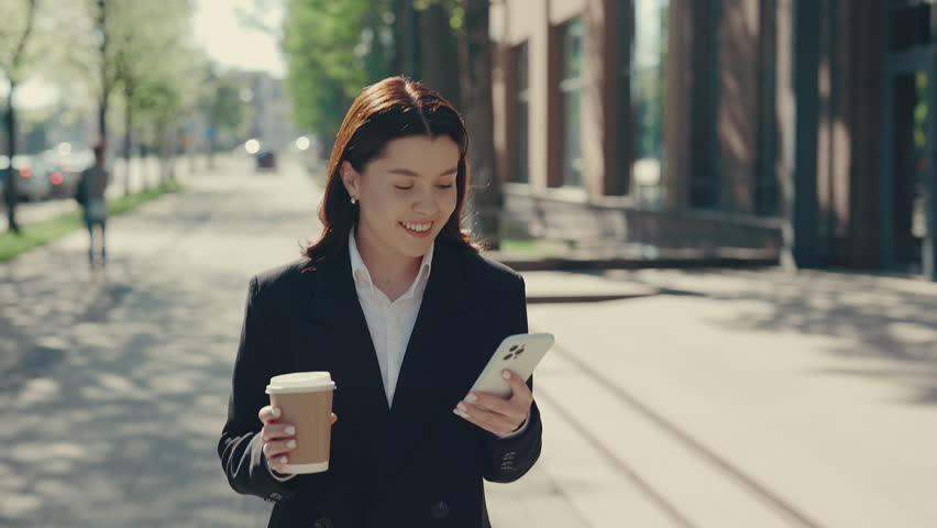 Attractive Female Manager Wearing Formal Suit Going to the Business Centre. Woman Holding Using the Mobile Phone, Texting with Clients, Swiping Apps and Holding Eco Cup for Coffee or Tea