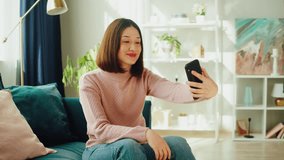 Asian smiling woman talking on video call. Young Korean female student speaking on smartphone, using virtual conference, sitting on sofa in living room. Communicating online via internet.