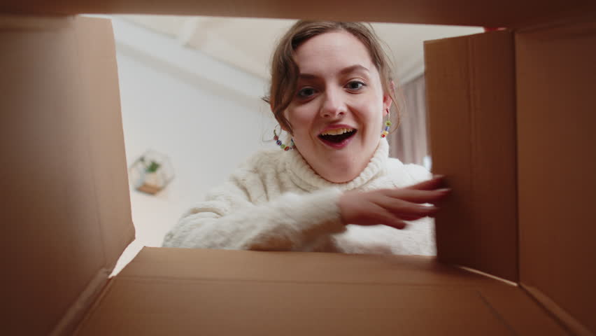 Inside view from box. Young happy woman unpacking delivery parcel at home. Smiling satisfied girl shopper, online shop customer opening cardboard receiving purchase gift by fast postal shipping order