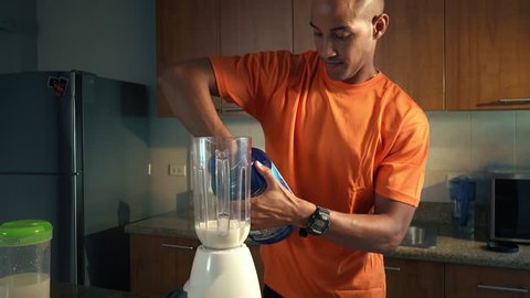 Young african american man in domestic kitchen, preparing a milk shake with proteins and vitamins. Black guy pours proteic supplement into the blender