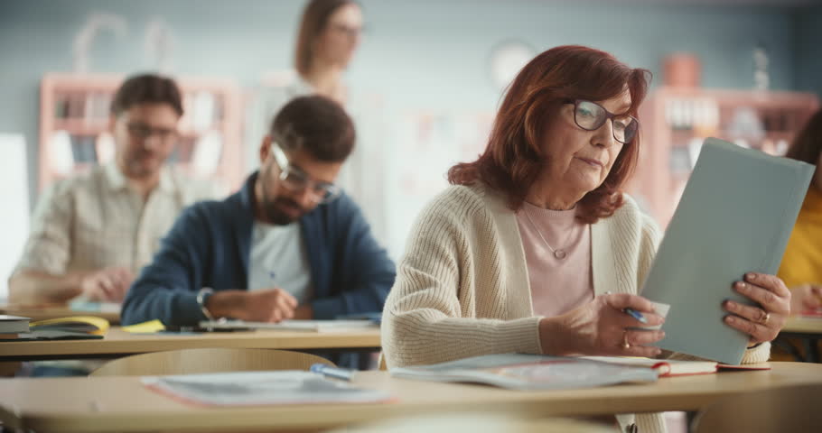Portrait of a Happy Senior Woman Taking a Course in an International Adult Education Center. Old Female Wearing Glasses, Sitting Behind a Desk, Using Textbook and Writing Down Notes in Notebook