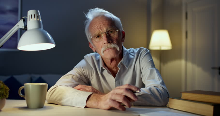 Adult old male psychologist drinks coffee during online video chat psychology session and looks at camera. Elderly man psychologist in semi-darkness of home office. Evening psychological session Royalty-Free Stock Footage #1103385849