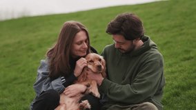 Young man and woman playing with cute dog outdoors. Couple and adorable puppy sitting on the grass