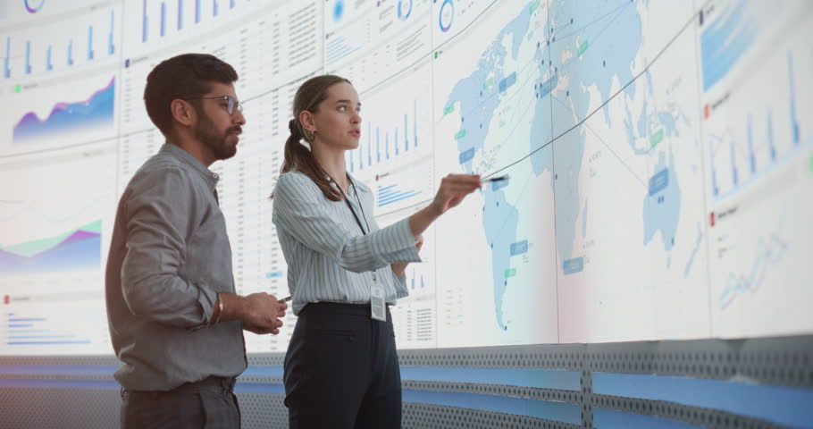 Caucasian Female Manager Talks To Indian Male Distribution Expert In Front Of Big Digital Screen With Infographics And Map In Monitoring Office. Successful Man And Woman Working In Logistics Company.